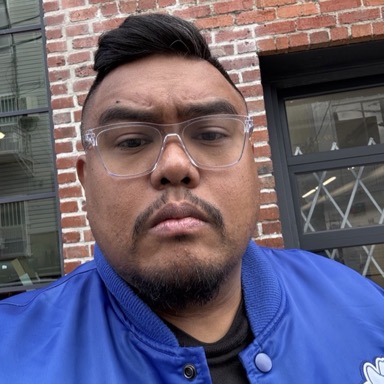 Kevin Dela Rosa in clear glasses and a blue jacket in front of a brick wall